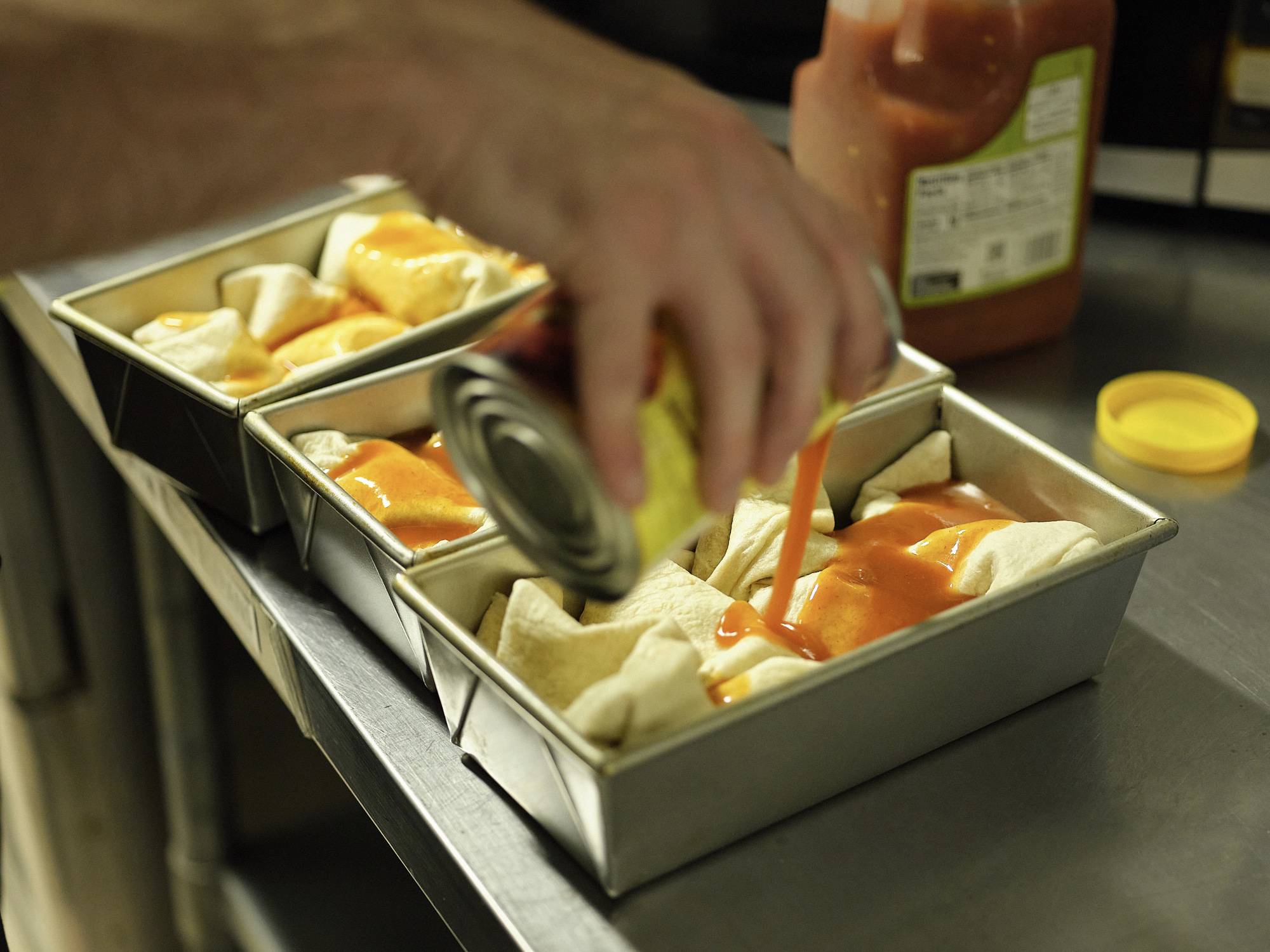 Scott making enchiladas. A hand pours a can of red sauce over taco shells in a bread pan